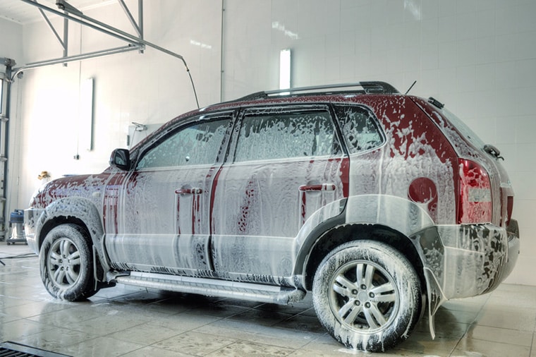 A red SUV is covered in white soap suds as it undergoes a wash in a brightly lit car wash facility. The car is positioned on a tiled floor, with overhead lights and a partially visible garage door in the background.