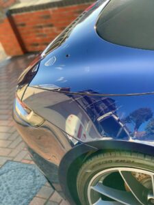 A close-up view of a sleek, reflective blue sports car parked on a brick driveway next to a brick wall. The car's rear quarter panel and tire are visible, with reflections of a house, trees, and pavement captured on the shiny surface.