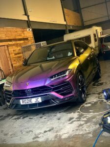 A metallic purple Lamborghini Urus SUV is parked inside a garage. The car's front grille and sleek design are prominently visible. The garage has wooden panels and other vehicles, including a caravan, in the background. The license plate reads "RO21E JC.