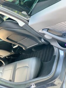 The image shows the interior of a car, specifically focusing on the rear seating area. The seats are upholstered in gray leather, and the car door is open, revealing part of the dashboard and steering wheel. The floor is covered with black striped carpet.