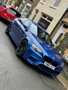 A shiny blue BMW car with the license plate "FM65 UCP" parked on a residential street beside a beige building with a white door and a green door. A red car is parked in front of the BMW, and the street is wet, indicating recent rain.