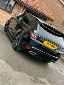 A black Range Rover SUV is parked on a driveway near a brick house with a tiled roof. The vehicle's license plate reads "J054 SHU." The car is shiny and well-maintained, with its rear view visible, showing its tail lights and exhaust pipes.