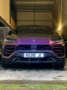 A sleek, purple Lamborghini Urus SUV is parked inside a dimly lit garage. The vehicle faces the viewer, showcasing its front grille and headlights. The license plate reads "RO21E JC." Fluorescent lights illuminate parts of the garage.