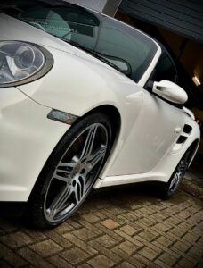 A close-up side view of a sleek, white sports car with low-profile tires and stylish alloy wheels parked on a brick driveway in front of a garage with a partially open door. The car's glossy finish reflects its surroundings.