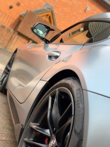 A close-up shot of a sleek, modern sports car with a matte finish, focusing on the rear wheel and part of the body. The Pirelli tire tread is visible, and the background features a brick building with a sloped roof.