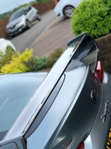 Close-up of the rear end of a grey sports car featuring a large black spoiler. Background shows a driveway with a few parked cars and green shrubs.