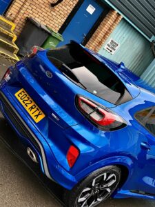 A blue Ford Puma car is parked outside a building with a brick wall and a blue door. The vehicle has a registration plate reading "EQ22 RTX" and is next to steps and a green trash bin. A sign on the wall reads "Helston Garages Motor Group.