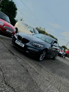 A sleek black BMW with a visible license plate "RF17 ZKD" is parked on the street, with several other cars, including a red Mini Cooper and a white van, in the background. Trees and a fence are also visible under a slightly cloudy sky.