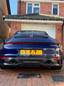 A blue Porsche sports car is parked in a driveway in front of a red-brick house with white trim. The car has a yellow license plate displaying the text "2 FJY." The house has white-framed windows and some Christmas lights hanging from the roof.