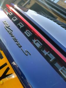 Close-up shot of the rear of a blue Porsche 911 Carrera S. The image focuses on the car's badge with the Porsche logo, "911 Carrera S" text, and part of a yellow license plate visible at the bottom left.
.