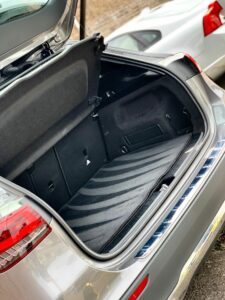 The image shows the trunk of an open hatchback car, with an empty, clean, black interior. The back seats of the car are visible, showing no items inside the trunk. There are a few other vehicles visible in the background on a street.