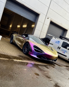 A sleek, iridescent sports car with a gradient finish ranging from gold to purple is parked outside a garage with its door partially open. To the right, a white van with roof equipment is parked near another garage door. The pavement is wet, indicating recent rain.