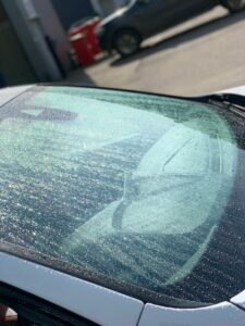 Close-up of a car's windshield covered in numerous water droplets, shining under direct sunlight. In the background, another car and a building are slightly out of focus, providing a blurred setting and context.