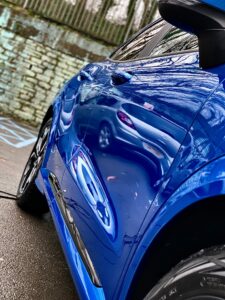 Close-up side view of a shiny blue car parked outdoors. The vehicle's surface vividly reflects its surroundings, including another car and trees. The ground is wet, and a stone wall with a fence at the top is visible in the background.