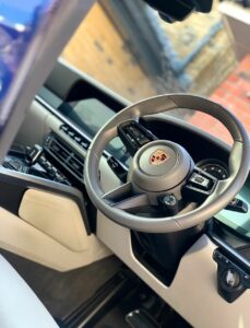 Interior of a modern car featuring a steering wheel with the Porsche logo in the center. The dashboard includes a digital display and various control buttons. The interior is primarily black and beige with some metallic accents visible.