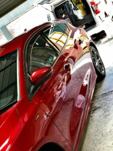 A shiny red car is parked in a garage. There is a white van in the background with its side door open, revealing cleaning supplies inside. The car's surface reflects the surroundings, showcasing the high-gloss finish.