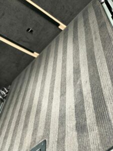 A close-up view of a carpeted floor with a stripe pattern consisting of alternating light and dark gray lines. The background shows parts of a metal frame and some structural elements.