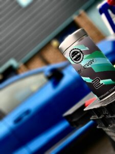 A close-up shot of a product labeled "SYNERGY 2" in a gray and teal design with the text "CERAMIC COAT." The background features a blurred blue car and a building, creating a focus on the product in the foreground.