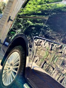 Close-up of a shiny black car, focusing on the left rear wheel and door. The vehicle's surface reflects greenery from the surrounding environment. The car is parked on a stone path with a wooden fence visible in the background.