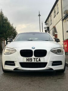 A white BMW parked on a residential street, captured from the front. The car has a license plate with the number "H19 TCA." The background includes a tree, houses, and a red recycling bin. The sky appears overcast.