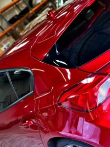 A close-up view of the rear side of a shiny red car, showcasing its sleek design and reflective surface. The car's rear window, taillight, and part of the rear door are visible. The background includes shelves with various items in a garage setting.