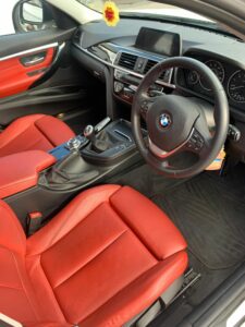 The interior of a car featuring red leather seats, a black dashboard, and a BMW emblem on the steering wheel. The car is equipped with a center console, various controls, a display screen, and an automatic gear stick. A hanging air freshener is visible.