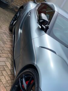 A sleek, silver sports car is parked on a brick driveway. The angle of the photo highlights the car's glossy finish, aerodynamic curves, and stylish black side mirror. The back wheel with a glimpse of red brake caliper is also visible.