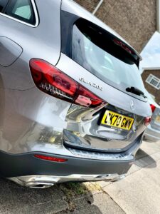 An image of the rear side of a silver Mercedes-Benz GLA 200 parked on the side of a street. The car has a yellow license plate with the alphanumeric sequence "LX70 GMW." A blurred background shows a portion of another vehicle and a building with brick walls.