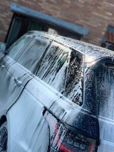 A black SUV is being washed, covered in white soap suds. The vehicle is parked in front of a brick building, and the sun is reflecting off the soapy surfaces, creating a bright glare on the car's windows and body.