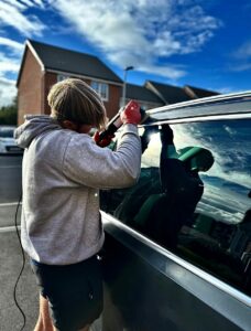 A person wearing a gray hoodie and red gloves is repairing or installing something on the side of a black car with a tool in hand. The car is parked in a parking lot with several houses visible in the background under a partly cloudy sky.