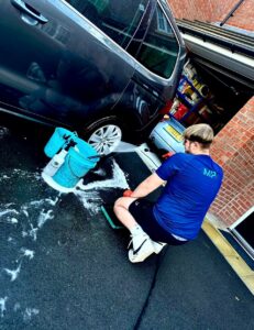 A person is washing a car in a driveway. They are using a hose to spray water on the car, with a blue bucket filled with water and suds nearby. The garage door is open, revealing various items stored inside. The person is wearing a blue shirt and black shorts.