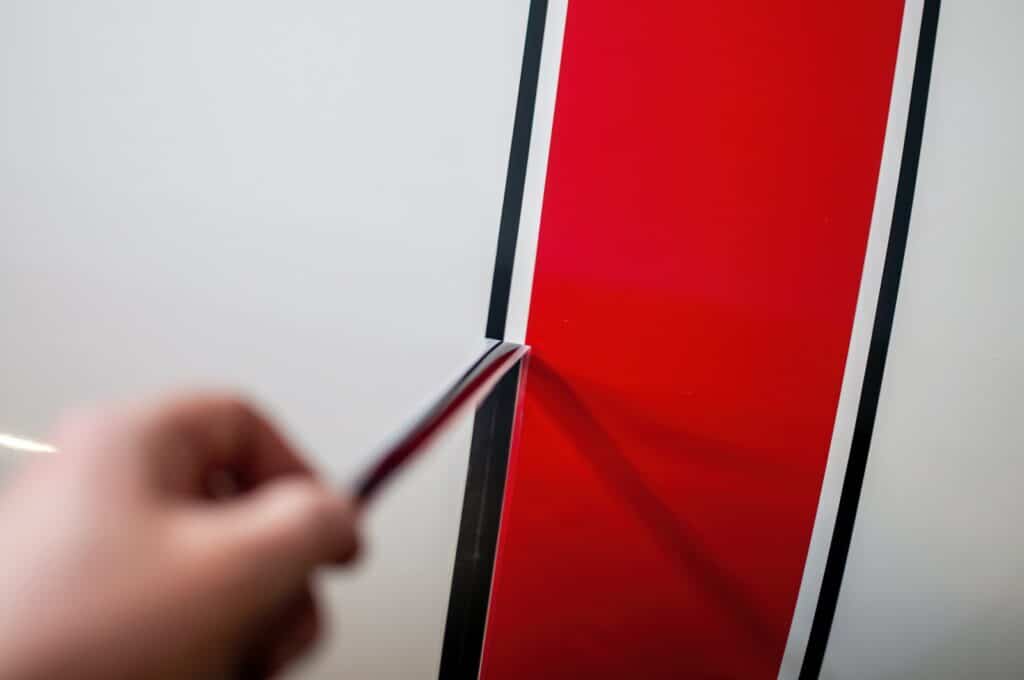 A blurred hand holds a cutting tool while applying a red stripe with black outlines on a smooth white surface.