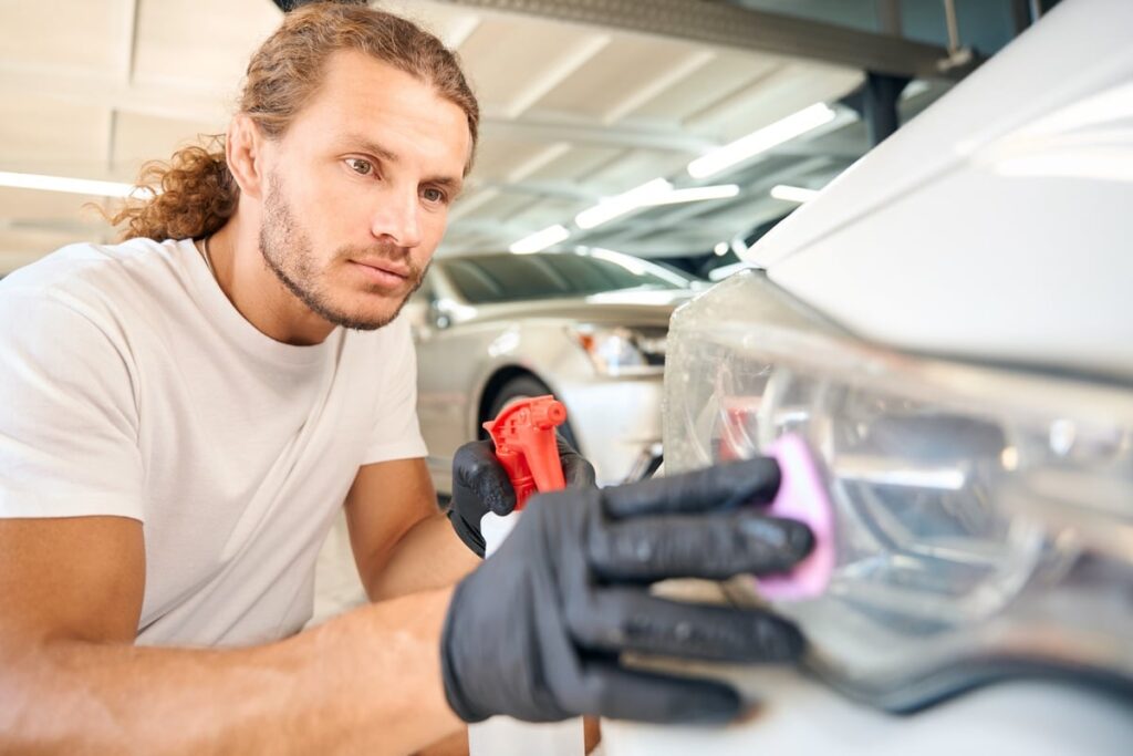 A man with long hair, wearing a white t-shirt and black gloves, polishes a car headlight in a brightly lit garage. He is focused on cleaning the surface with a pink cloth, and a spray bottle is nearby.