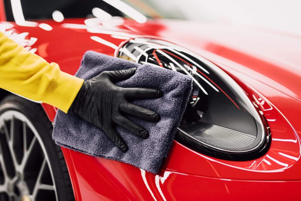 A person wearing a yellow shirt and black gloves polishes the headlight of a shiny red sports car with a gray towel. The close-up image focuses on the hand, towel, and headlight.